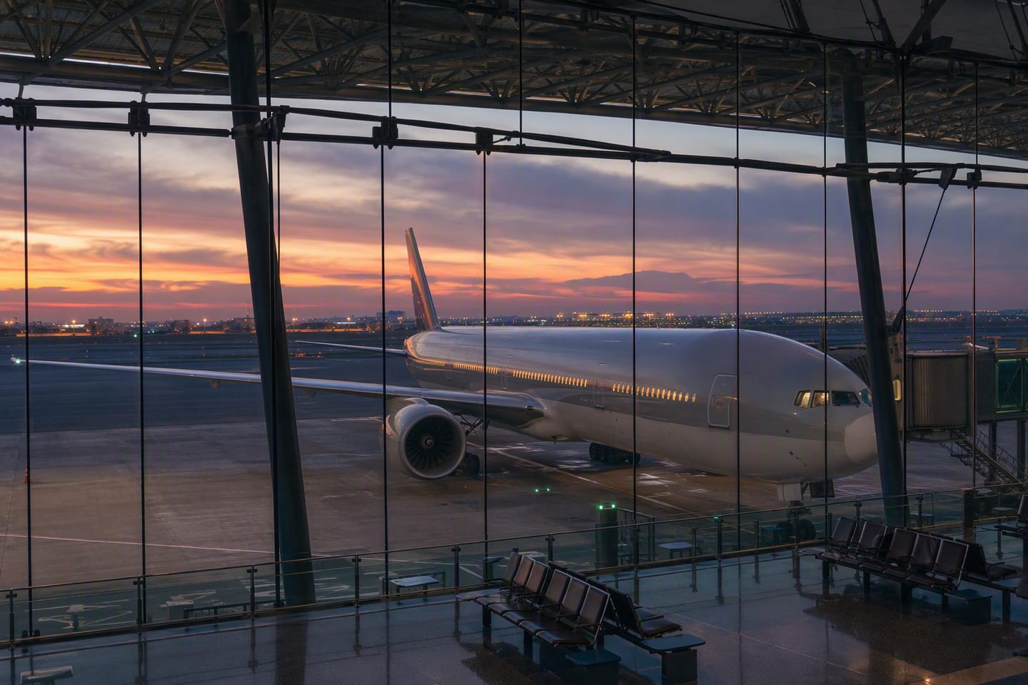 A Qatar Airways Boeing 777 sits at a quiet gate beneath the glass roof of Hamad International Airport in Doha at dusk, its cabin lights glowing against the empty terminal.