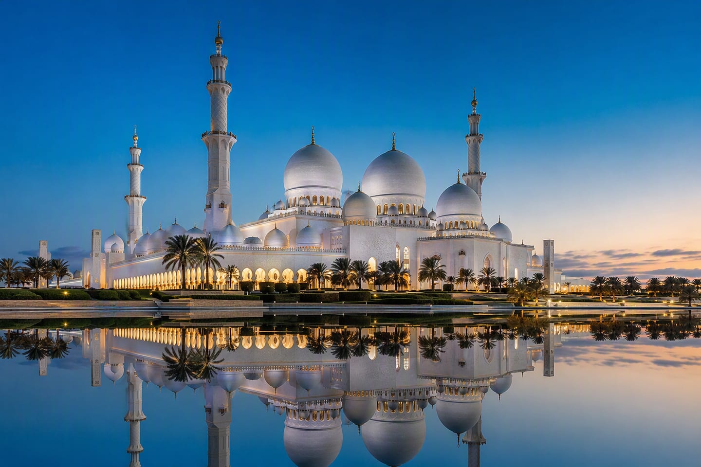 Abu Dhabi’s Sheikh Zayed Grand Mosque glows white at dusk beneath a deepening blue sky, its domes and minarets reflected in the still surrounding pools.