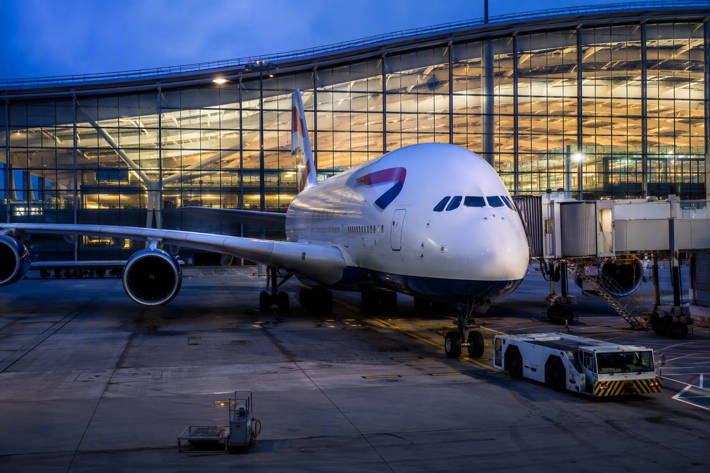 A British Airways Airbus A380 sits at the gate beneath the illuminated glass roof of London Heathrow’s Terminal 5 at blue hour.