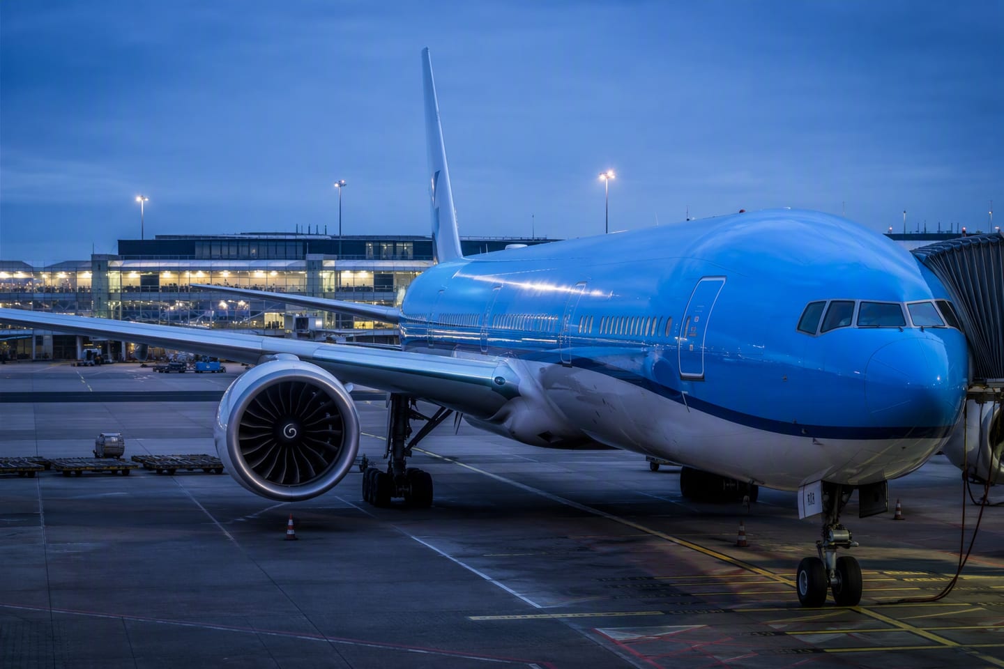 A KLM Boeing 777 sits at a quiet gate beneath the blue-hour lights of Amsterdam Airport Schiphol, its polished fuselage reflecting the terminal windows and empty tarmac.