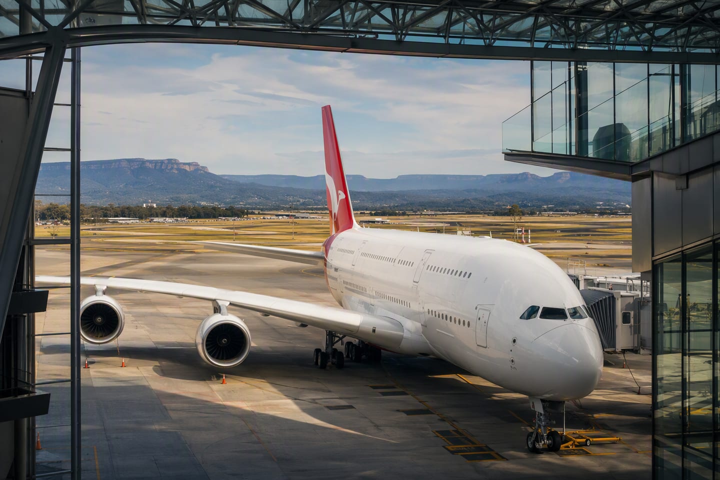 A Qantas Airbus A380 rests at the gate beneath the glass terminal canopy at Sydney Kingsford Smith Airport, with the Blue Mountains faintly visible beyond the runway.