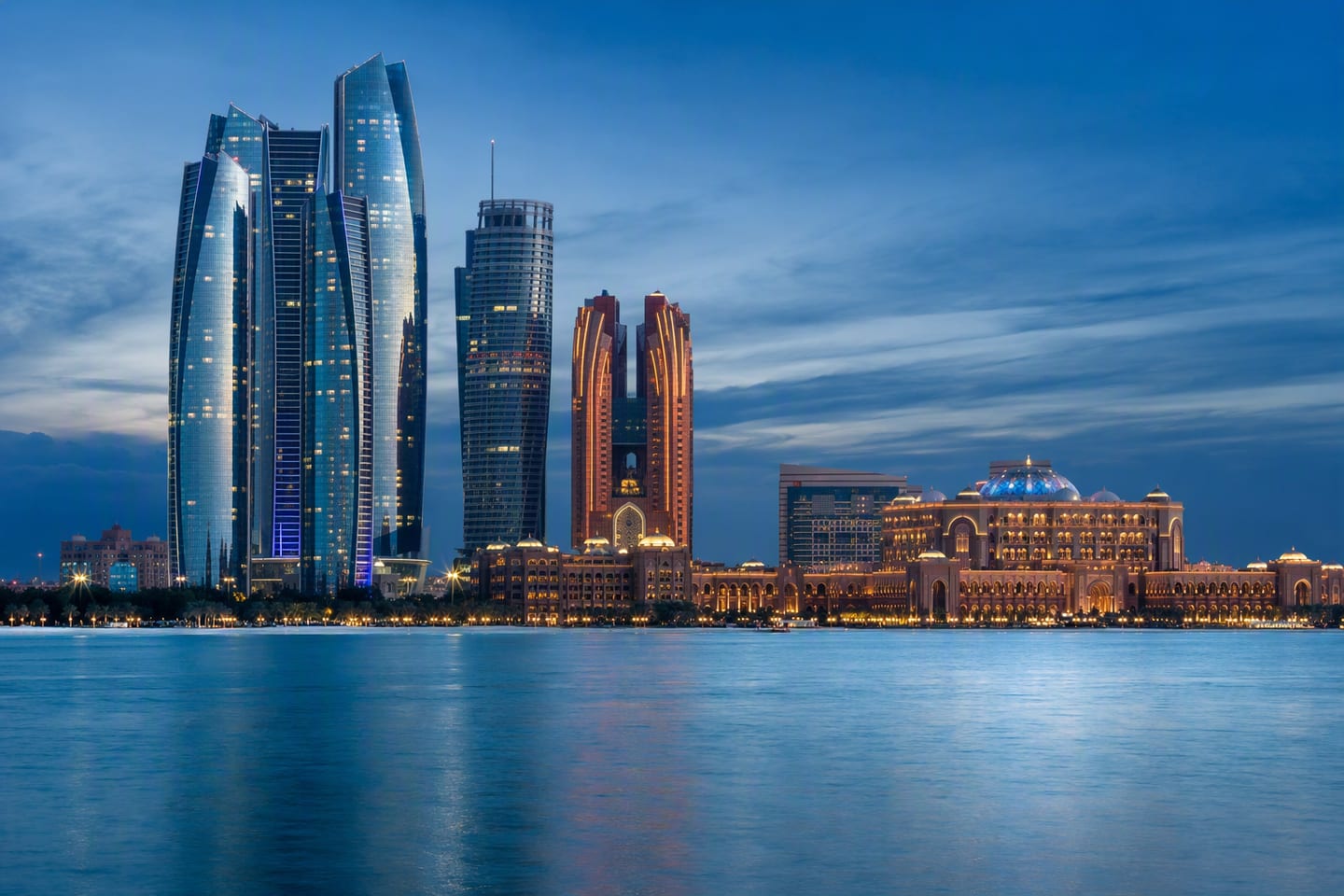 The Abu Dhabi skyline rises beyond the Corniche at blue hour, with the Etihad Towers and Emirates Palace glowing above the calm Gulf.