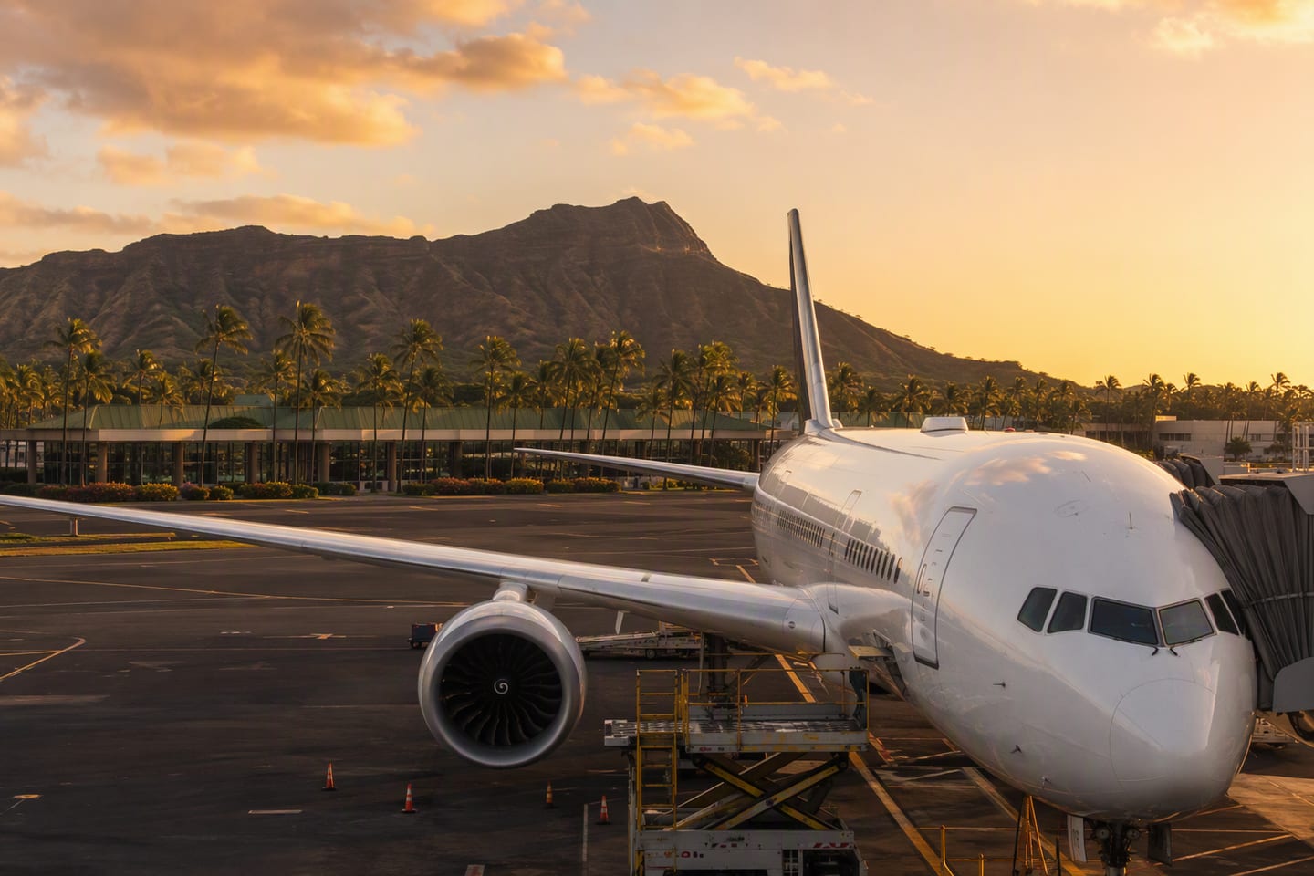 A wide-body jet stands at the gate at Honolulu’s Daniel K. Inouye International Airport at golden hour, with Diamond Head rising beyond the palm-lined terminal.