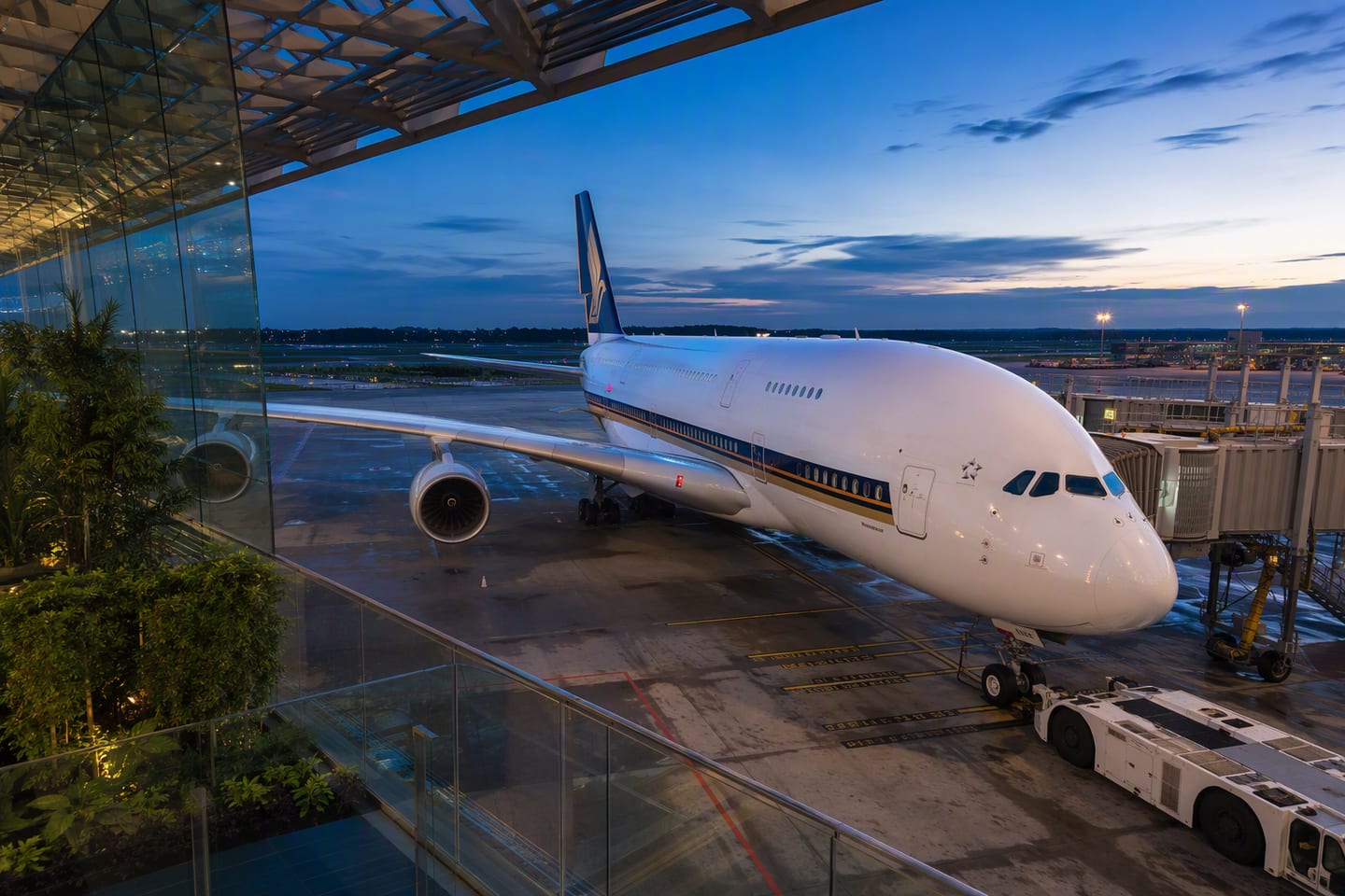 A Singapore Airlines Airbus A380 rests at the gate beneath the glass roof of Singapore Changi Airport, its polished fuselage glowing in the blue light of early evening.
