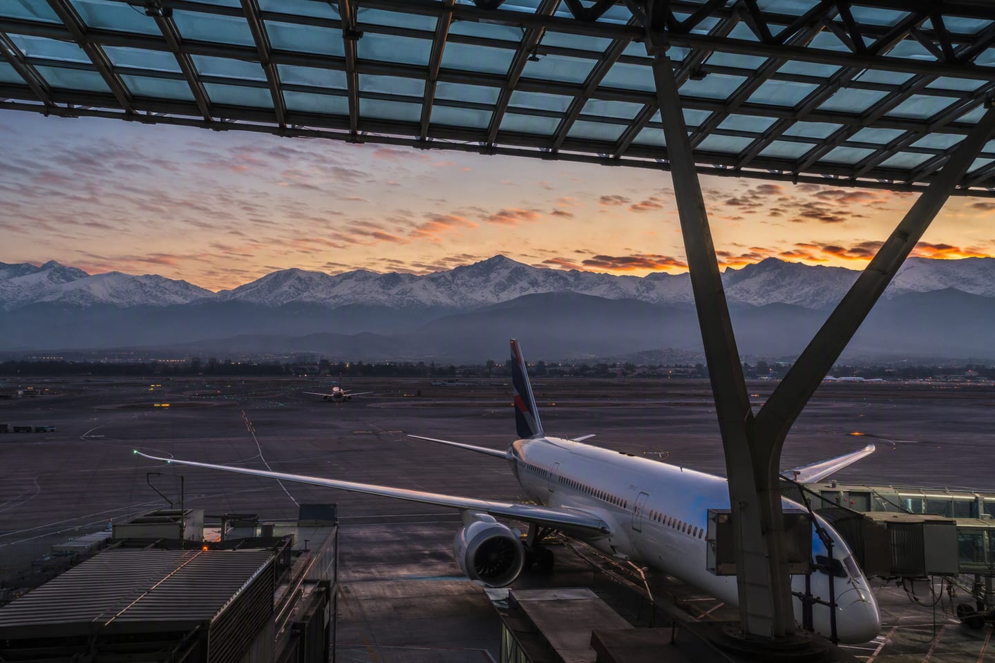 A LATAM Boeing 787 Dreamliner rests at a gate beneath the glass roof of Santiago International Airport, with the Andes rising beyond the terminal at dusk.