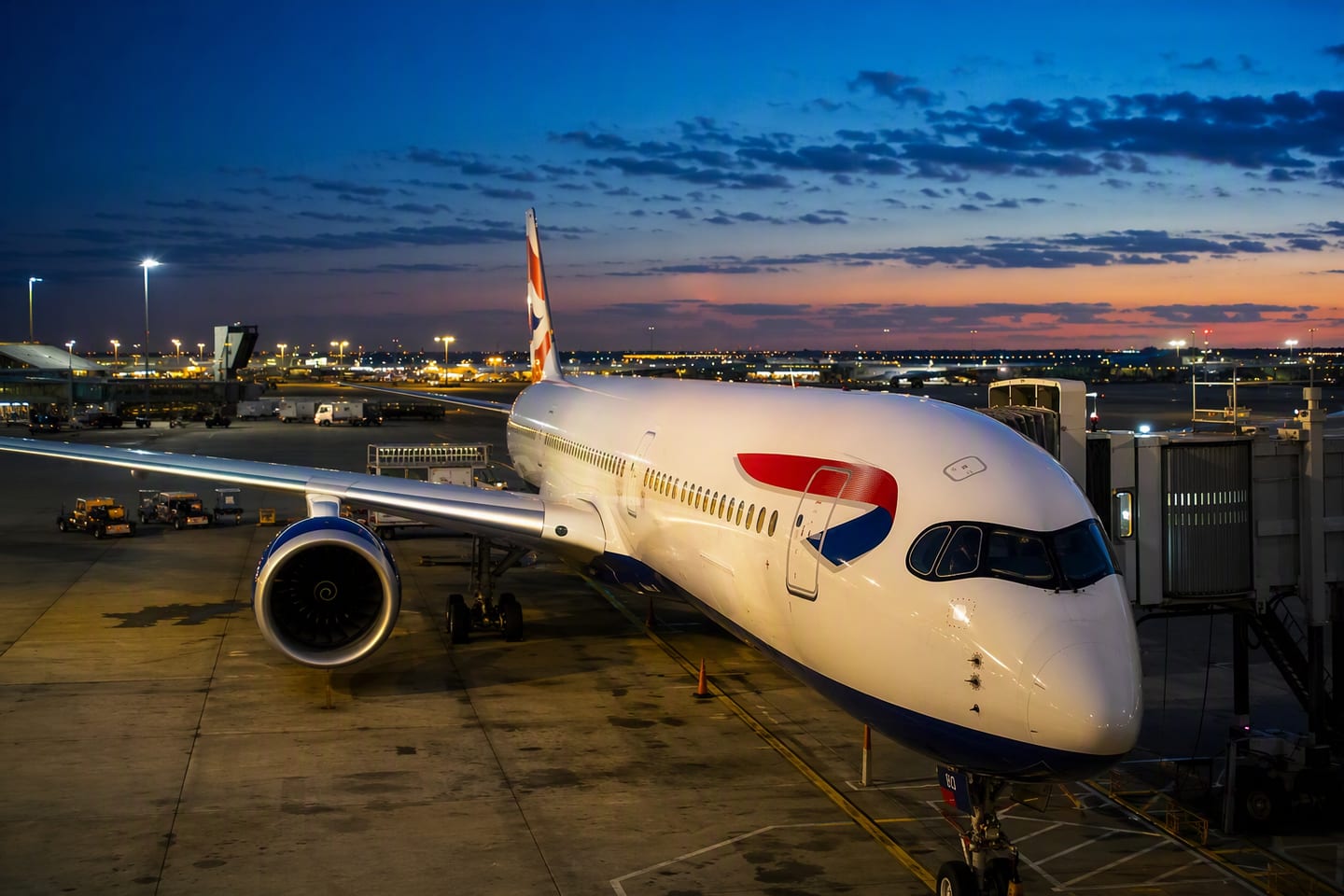 A British Airways Airbus A350-1000 sits at the gate at New York JFK’s Terminal 8 at dusk, with the airport lights glowing beneath a deep blue sky.