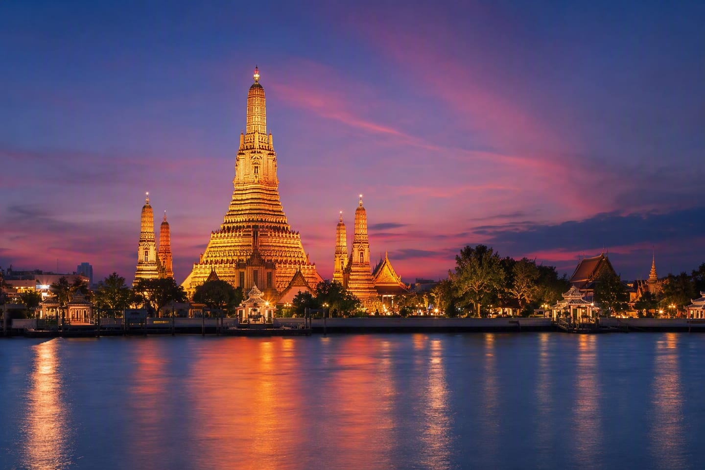 Bangkok’s Wat Arun rises beside the Chao Phraya River at blue hour, its illuminated porcelain spires mirrored in the calm water beneath a deepening violet sky.