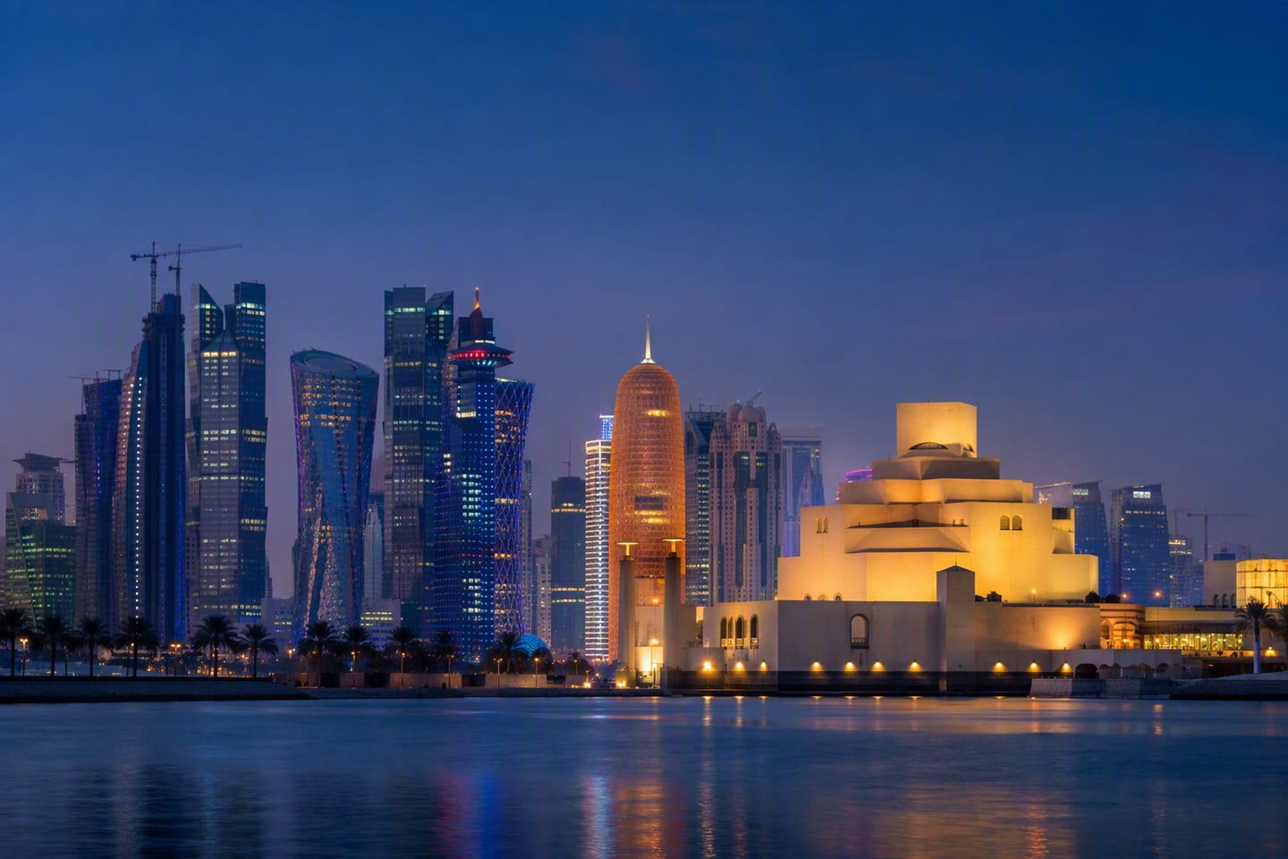 Doha’s illuminated West Bay skyline rises behind the calm Corniche at blue hour, with the Museum of Islamic Art silhouetted in the foreground and no people visible.