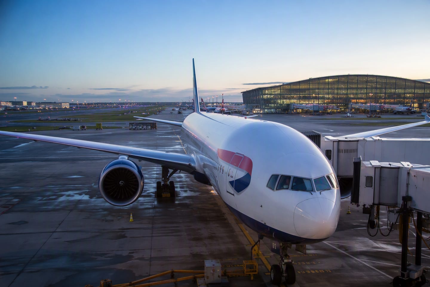 A British Airways Boeing 777 sits at a Terminal 5 gate at London Heathrow beneath the soft blue light of dawn, with the glass terminal and runway stretching behind it.