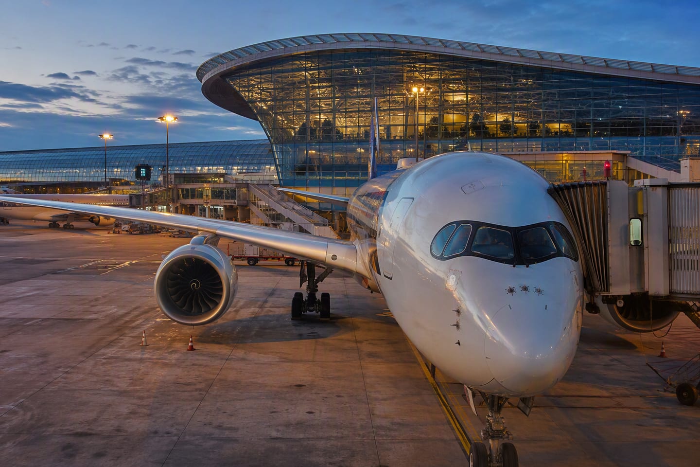 A Singapore Airlines Airbus A350 sits at a gate beneath the sweeping glass roof of Singapore Changi Airport, its polished fuselage glowing in the blue-hour light.